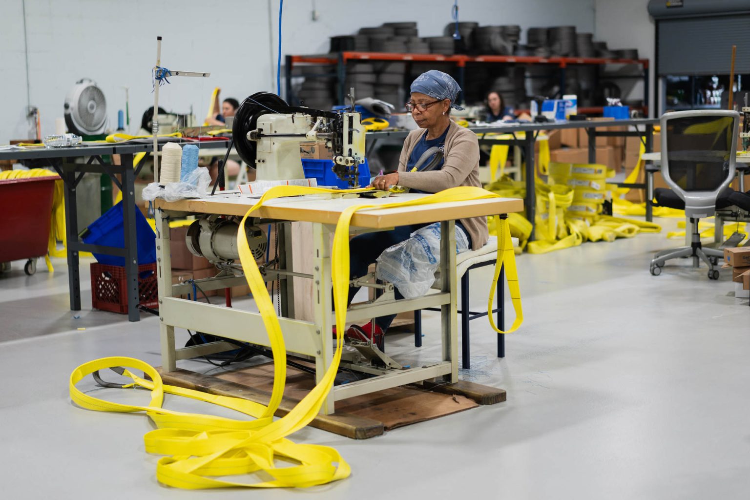 At the sewing machine, working on sewing yellow slings for shipment with other team members in the background