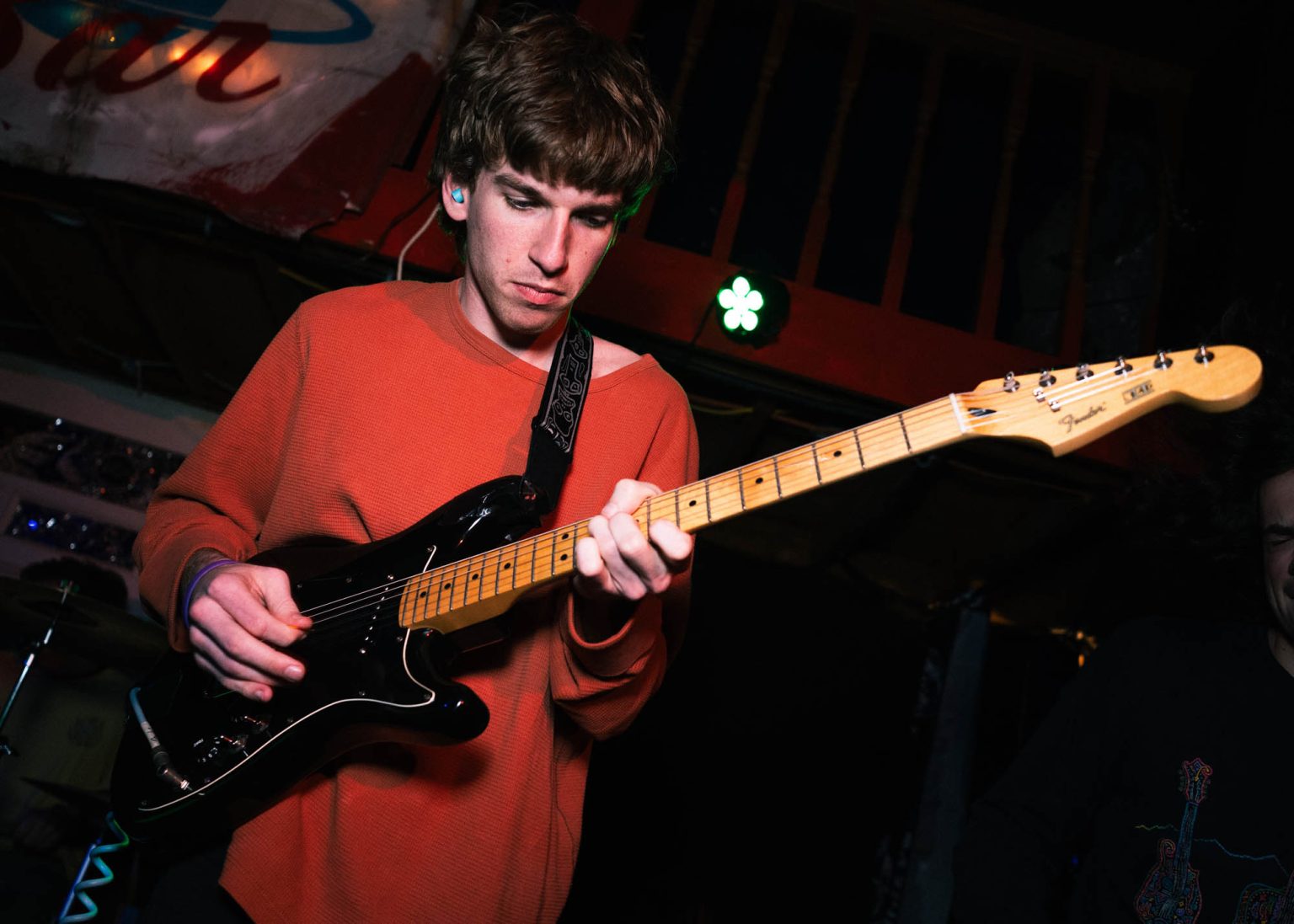 Guitarist on stage focused on playing his guitar in the background on the left and right of him we see fellow band mates