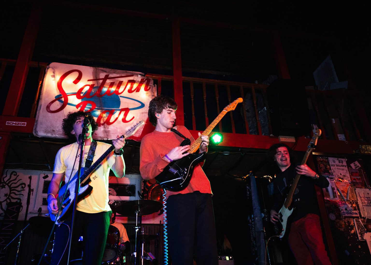 Lead singer, guitarist, and bassist on stage during their performance with the sign of the venue in the background Saturn Bar