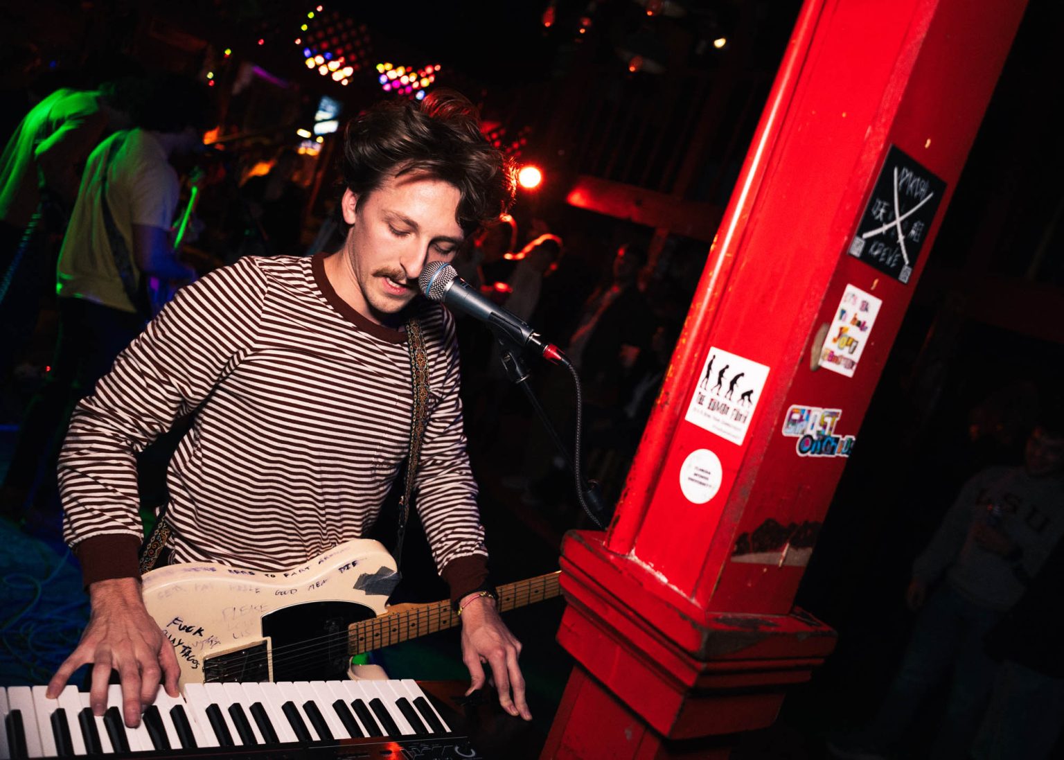 Guitarist with his guitar not playing that instrument but instead playing the keyboard during their live performance at a local bar with crowd and fellow band mates in the background