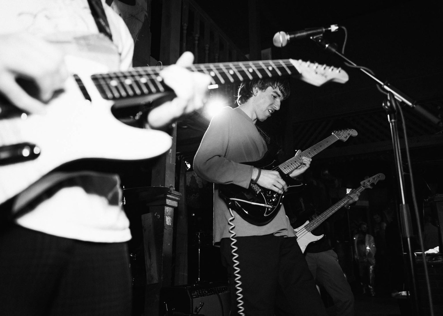 Guitar in the foreground and lead guitarist smiling in the mid-ground and bassist in the background along with some crowd at the live performance