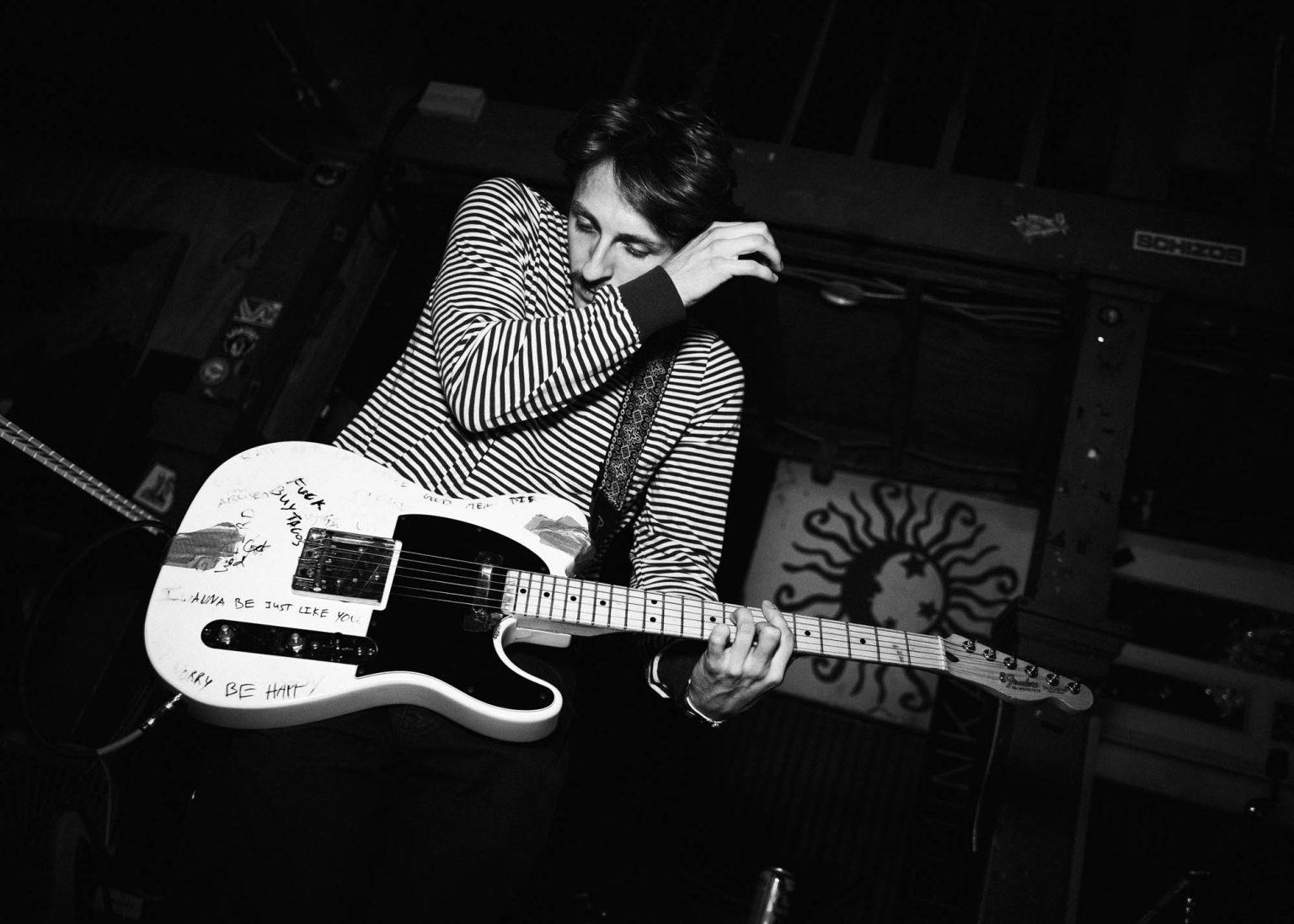 Guitarist in striped black and white shirt is covering face with his arm while playing guitar on stage during their live performance