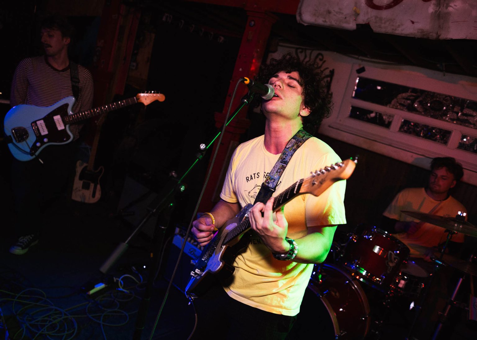 Lead singer singing into microphone while playing guitar along with fellow guitarist and drummer in the background