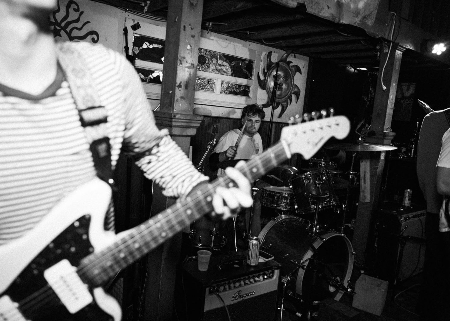 Guitarist in the foreground playing guitar with drummer in the background during concert