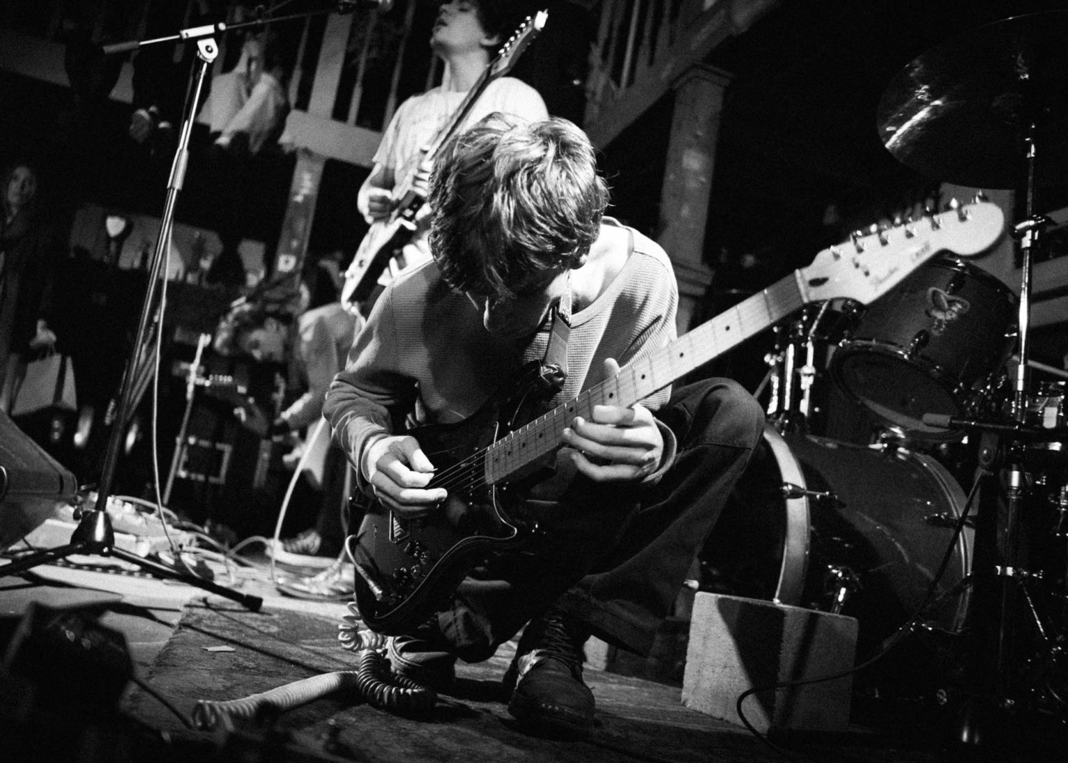 Guitarist crouched down playing guitar with fellow band mates in the backgorund